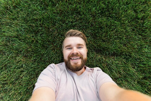 Joven con barba y peinado de moda tirado en un césped tomando selfie sosteniendo un teléfono inteligente o una tableta y mirando a la cámara