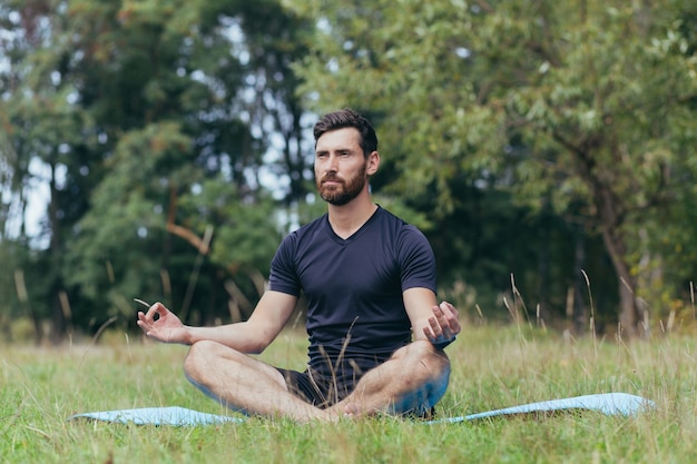 Un joven con barba sentado en el parque sobre una estera medita, realiza ejercicios para mejorar la respiración, estilo de vida activo