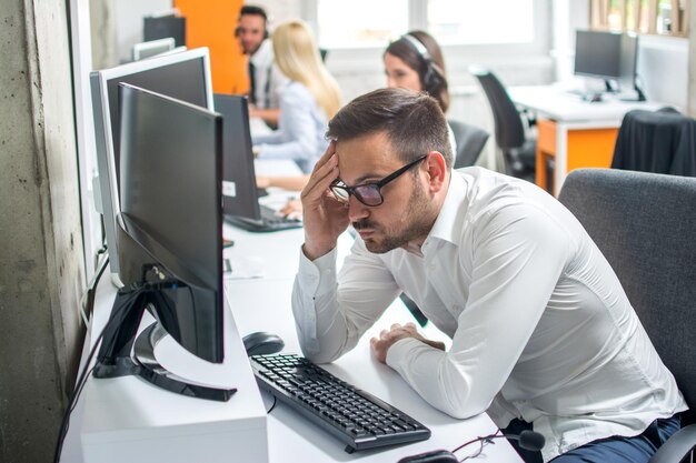 Un joven con barba serio mirando la pantalla de la computadora mientras está sentado en su lugar de trabajo en la oficina