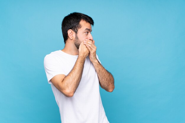 Joven con barba sobre azul aislado cubriendo la boca y mirando hacia un lado