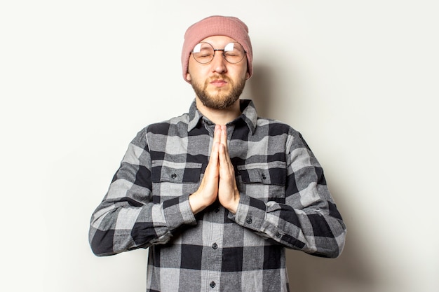 Foto joven con barba en un sombrero, camisa a cuadros y gafas está meditando o rezando en un blanco aislado. gesto de meditación, relájate