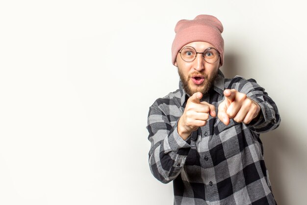 Joven con una barba en un sombrero, una camisa a cuadros señala con sus dedos al espectador en un blanco aislado. El gesto que eres el próximo, te eligió a ti.