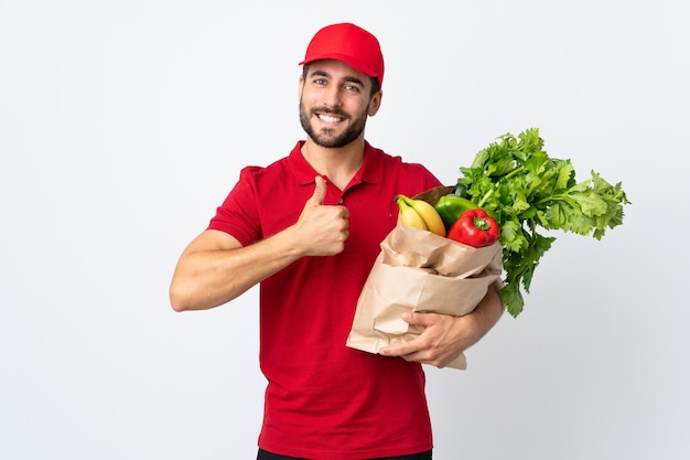 Joven con barba sosteniendo una bolsa llena de verduras aisladas en la pared blanca dando un gesto de pulgares arriba