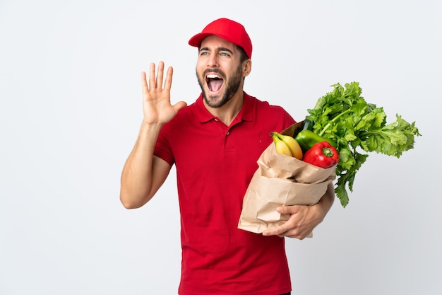 Joven con barba sosteniendo una bolsa llena de verduras aisladas en la pared blanca gritando con la boca abierta