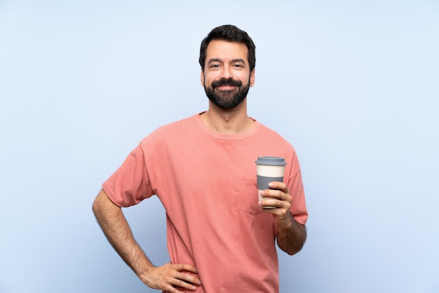 Joven con barba sosteniendo un café para llevar sobre azul aislado posando con los brazos en la cadera y sonriendo