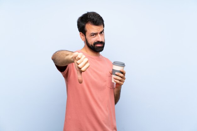 Joven con barba sosteniendo un café para llevar sobre la pared azul aislado mostrando el pulgar hacia abajo con expresión negativa