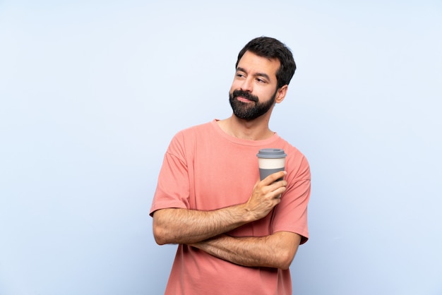 Joven con barba sosteniendo un café para llevar sobre retrato de pared azul aislado