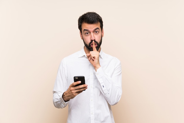 Joven con barba sosteniendo un móvil mostrando un gesto de silencio poniendo el dedo en la boca
