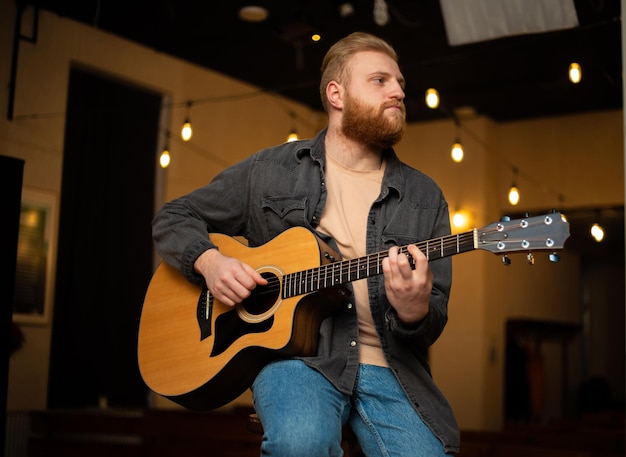 Un joven con barba toca una guitarra acústica en una habitación con una iluminación cálida