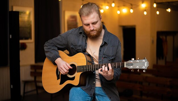 Un joven con barba toca una guitarra acústica en una habitación con una iluminación cálida