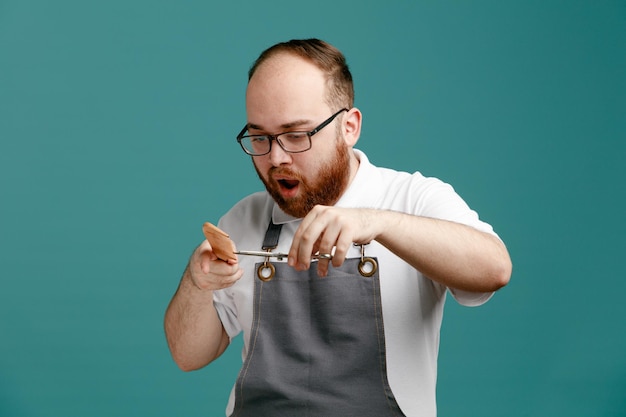 Un joven barbero impresionado con uniforme y gafas mirando hacia abajo sosteniendo un peine y unas tijeras finge hacer un corte de pelo para un cliente aislado de fondo azul