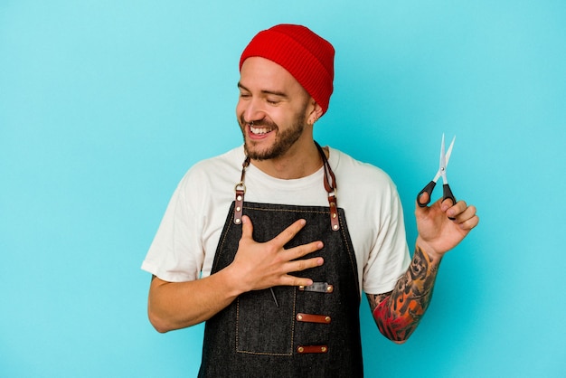 Joven barbero tatuado aislado sobre fondo azul se ríe a carcajadas manteniendo la mano en el pecho.