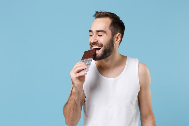 Joven barbudo de 20 años con camisa blanca sostiene una barra de chocolate en la mano aislada en un fondo de pared azul pastel, retrato de estudio. Concepto de procedimientos cosméticos para el cuidado de la piel. Simulacros de espacio de copia