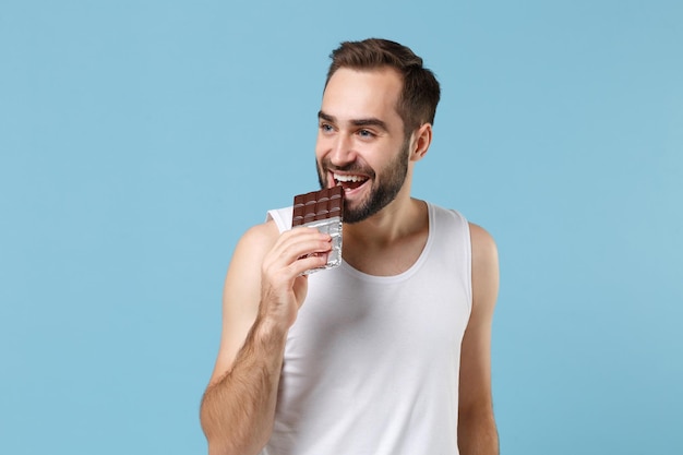 Joven barbudo de 20 años con camisa blanca sostiene una barra de chocolate en la mano aislada en un fondo de pared azul pastel, retrato de estudio. Concepto de procedimientos cosméticos para el cuidado de la piel. Simulacros de espacio de copia