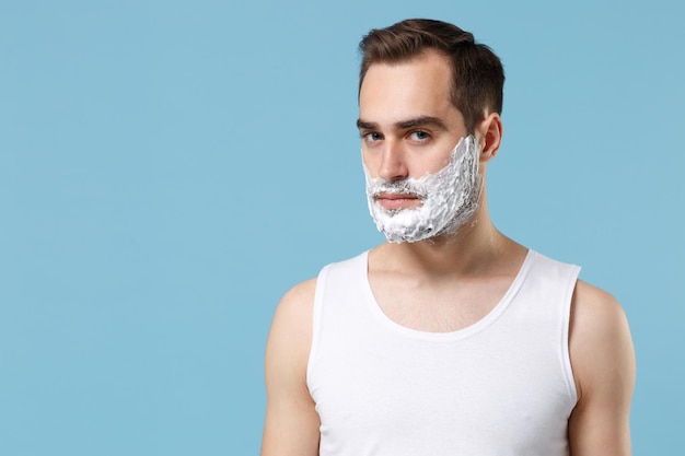 Joven barbudo de 20 años con la cara de camisa blanca cubierta con espuma de afeitar aislada en un retrato de estudio de fondo azul pastel. Concepto de procedimientos cosméticos para el cuidado de la piel. Simulacros de espacio de copia.