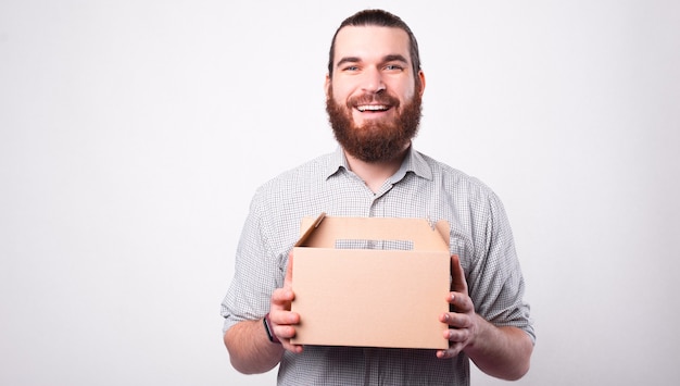 Un joven barbudo alegre está sosteniendo una caja de entrega y sonriendo está mirando a la cámara cerca de una pared blanca