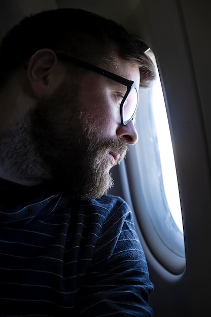 Joven barbudo con anteojos mira pensativo y triste por la ventana de un avión