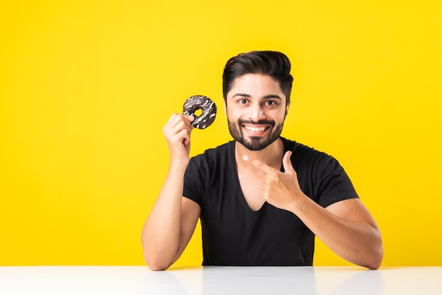 Joven barbudo asiático indio come donut o donut mientras está sentado en la mesa de la cocina contra el fondo amarillo o blanco