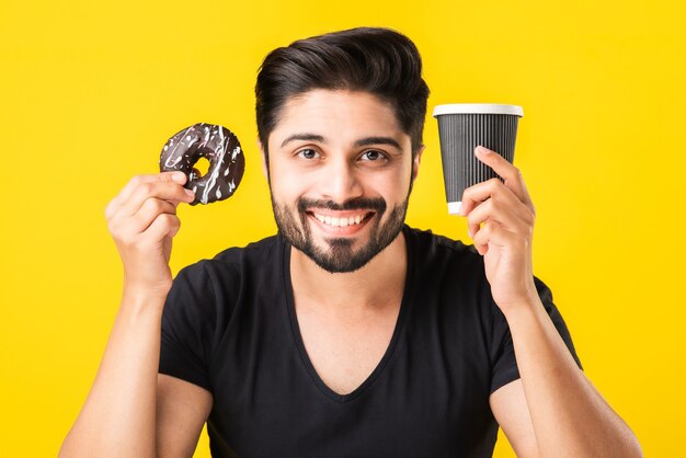 Joven barbudo asiático indio come donut o donut mientras está sentado en la mesa de la cocina contra el fondo amarillo o blanco