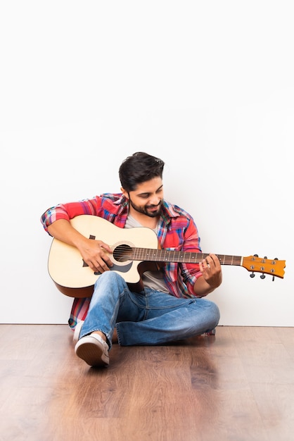 Foto joven barbudo asiático indio tocando la guitarra acústica mientras está sentado aislado sobre un piso de madera contra la pared blanca