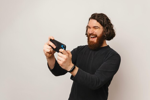 Foto un joven barbudo asombrado jugando juegos en línea en su teléfono inteligente sobre un fondo blanco