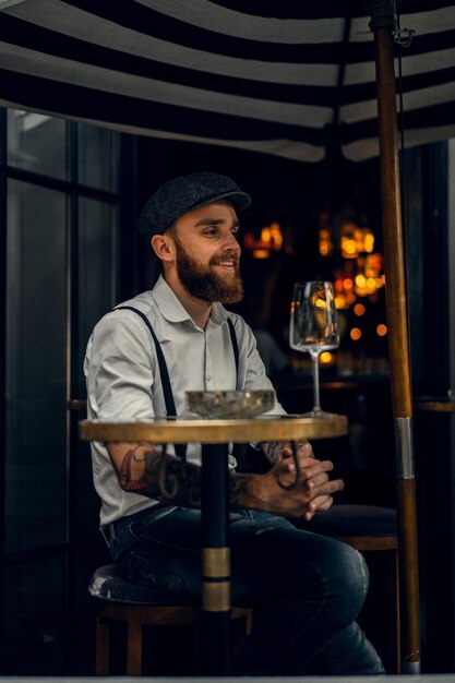 Joven barbudo en un café en la calle con una copa de vino. Chico romántico con una gorra de camisa blanca y tirantes en la ciudad. Peaky Blinders. antigua moda retro.