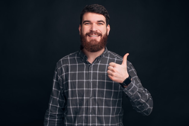 Joven barbudo con una camisa, está mostrando como o pulgar arriba gesto en la pared negra