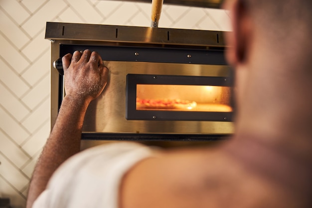 Joven barbudo cocinando en un restaurante y haciendo pizza mientras usa el horno