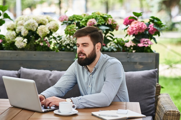 Joven barbudo con una computadora portátil en el café al aire libre