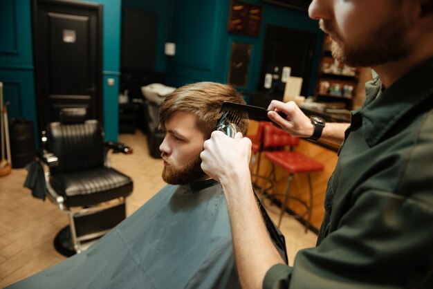 Joven barbudo cortarse el pelo por peluquero con maquinilla de afeitar eléctrica mientras está sentado en una silla