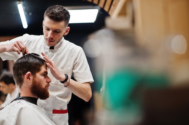 Joven barbudo corte de pelo por peluquero mientras está sentado en la silla en la barbería. Alma de barbero.