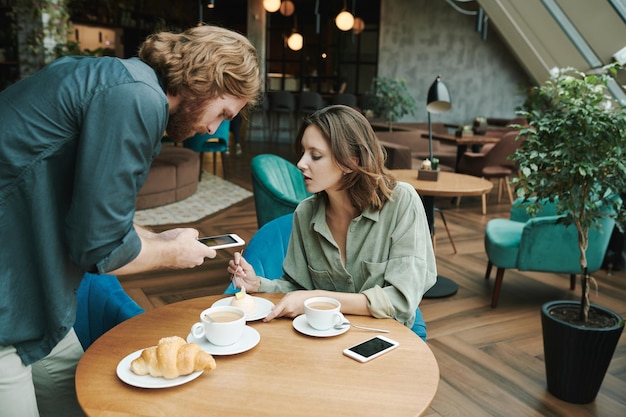 Un joven barbudo disgustado sentado en un café moderno y esperando una respuesta mientras llama por celular