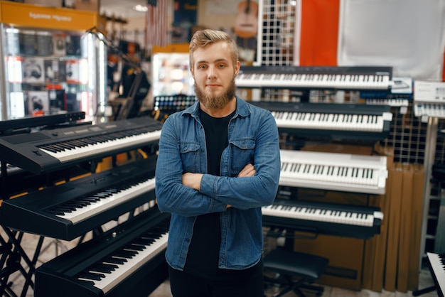 Joven barbudo eligiendo sintetizador en la tienda de música. Surtido en la tienda de instrumentos musicales, músico masculino comprando equipos