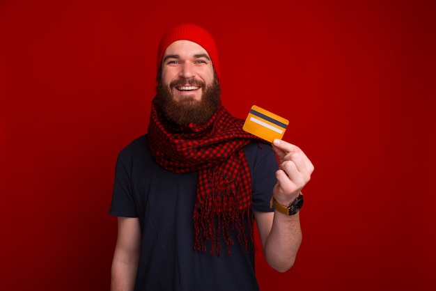 Joven barbudo está sonriendo y mostrando una tarjeta de crédito cerca de fondo rojo.