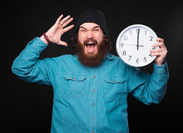 Un joven barbudo estresado sostiene un gran reloj blanco y se ve muy estresado ante la cámara cerca de una pared negra