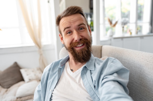 Joven barbudo feliz sonriendo y tomando una foto selfie mientras se sienta en el sofá en casa