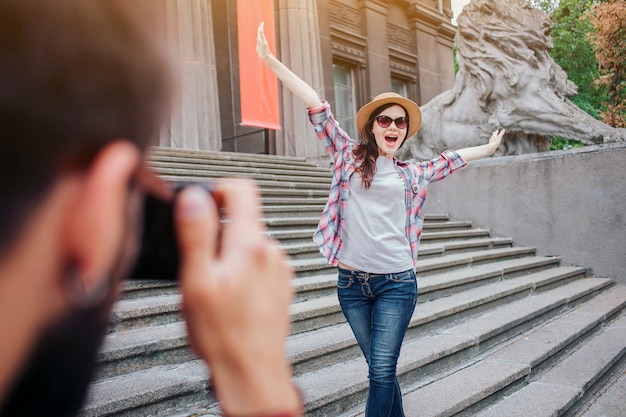 Joven barbudo hablando imagen de mujer feliz. Ella posa ante la cámara. Turista mantenga las manos en alto y usa gafas. Se paran en las escaleras