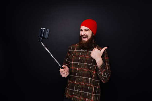 Joven barbudo haciendo un selfie con un palo selfie y mostrando el pulgar hacia arriba.