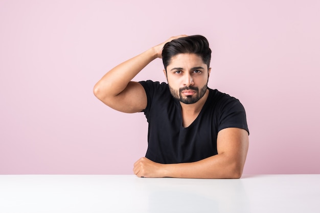 Joven barbudo indio pensando mientras está sentado aislado contra el fondo de color rosa en la mesa o escritorio