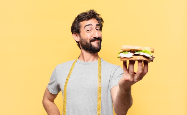 Joven barbudo loco haciendo dieta expresión feliz y sosteniendo un sándwich