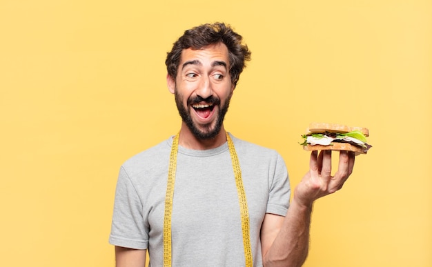 Joven barbudo loco haciendo dieta expresión de sorpresa y sosteniendo un sándwich