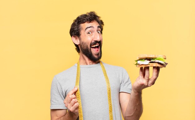 Joven barbudo loco haciendo dieta expresión de sorpresa y sosteniendo un sándwich