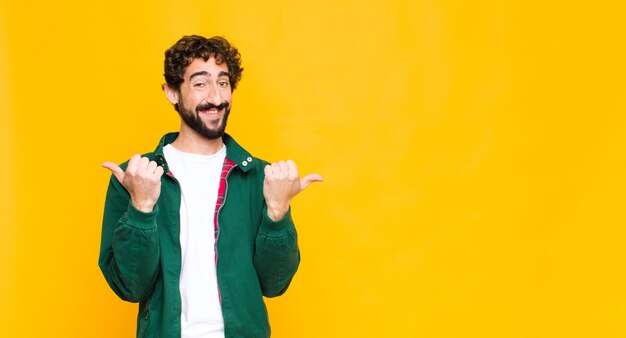 Joven barbudo loco sonriendo alegremente y luciendo feliz, sintiéndose despreocupado y positivo con ambos pulgares contra la pared plana