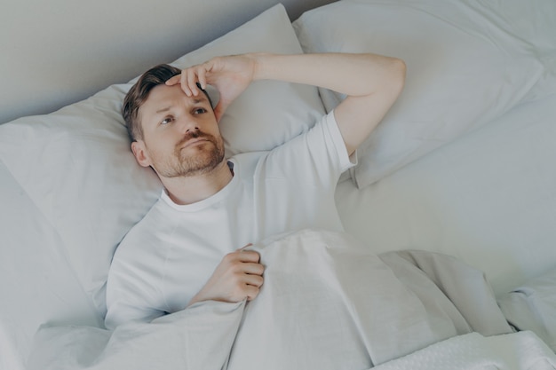 Joven barbudo molesto y cansado acostado en la cama con los ojos abiertos y no puede dormir, el hombre tiene problemas de insomnio o problemas para dormir. Concepto de dificultad para dormir y descansar