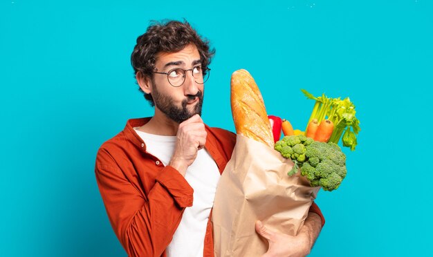 Joven barbudo pensando, sintiéndose dudoso y confundido, con diferentes opciones, preguntándose qué decisión tomar y sosteniendo una bolsa de verduras