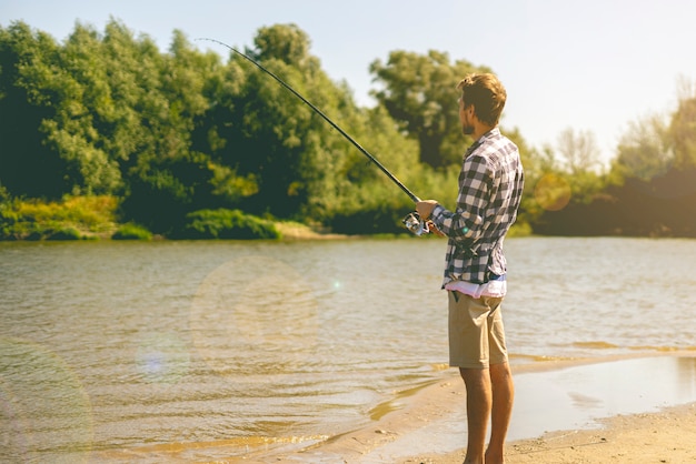 Joven barbudo pesca de pie en la orilla del río de arena con caña de pescar durante el verano.