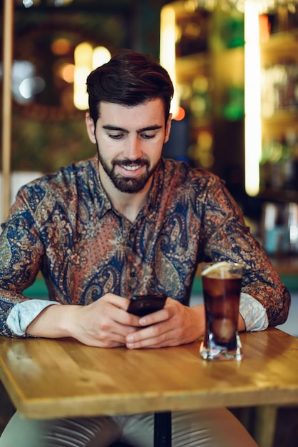 Joven barbudo con ropa casual mirando su teléfono inteligente en un pub moderno