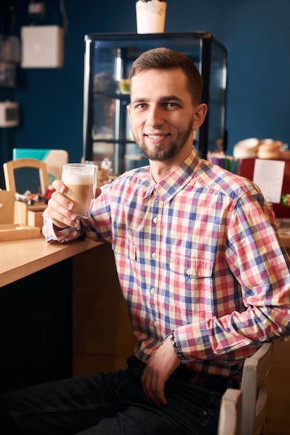 Joven barbudo sentado en un café y sonriendo a la cámara sosteniendo una taza de café con leche Fotografía de retrato Fondo oscuro