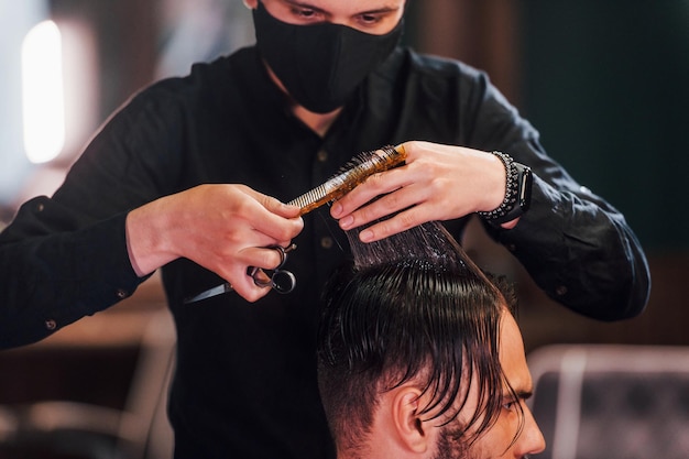 Joven barbudo sentado y cortándose el pelo en la peluquería por un tipo con máscara protectora negra