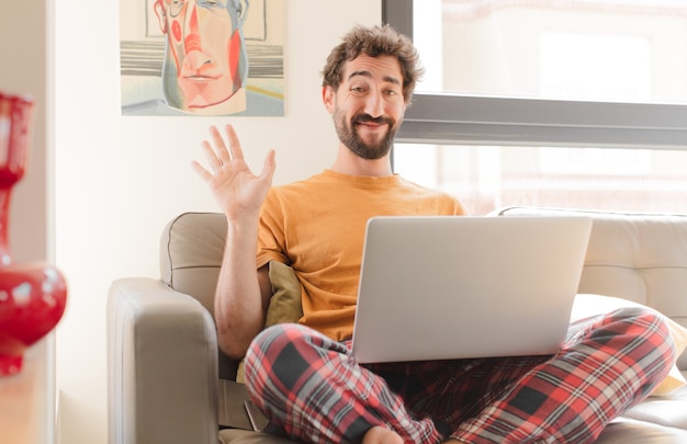 Joven barbudo sonriendo feliz y alegremente agitando la mano y saludándote o diciéndote adiós y sentado con una computadora portátil